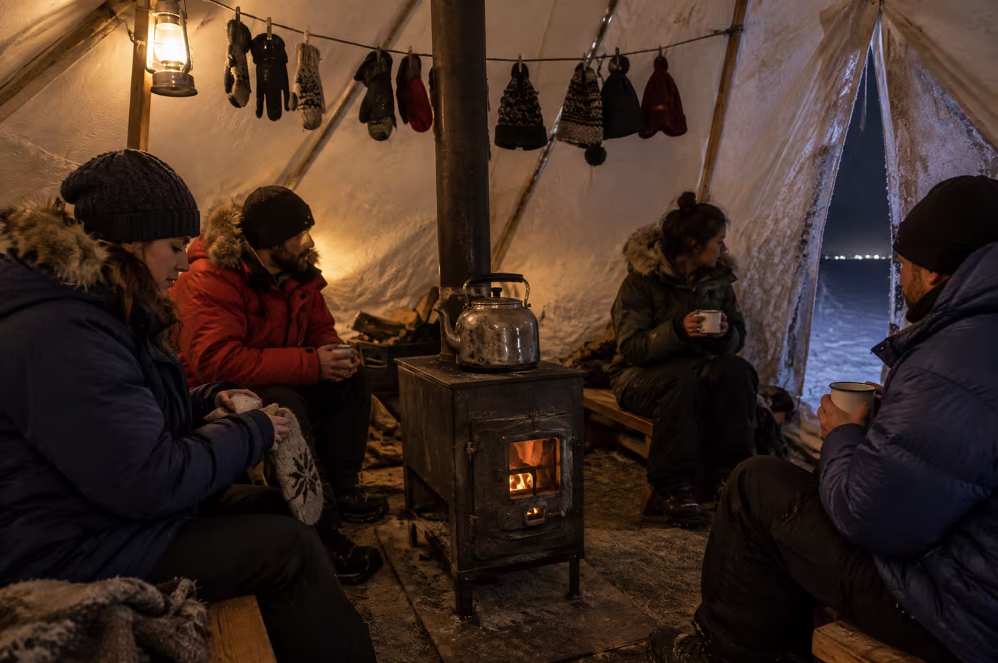 Guests in parkas drinking hot chocolate around a wood stove inside a canvas wall tent