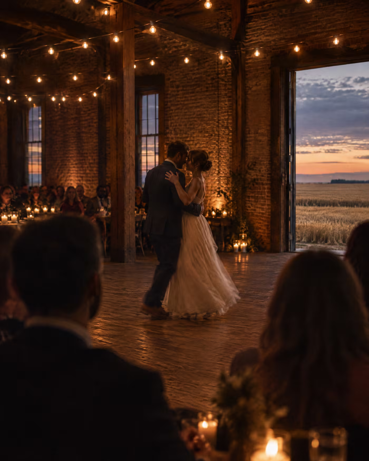A couple's first dance under strings of bare bulbs, guests watching in silhouette