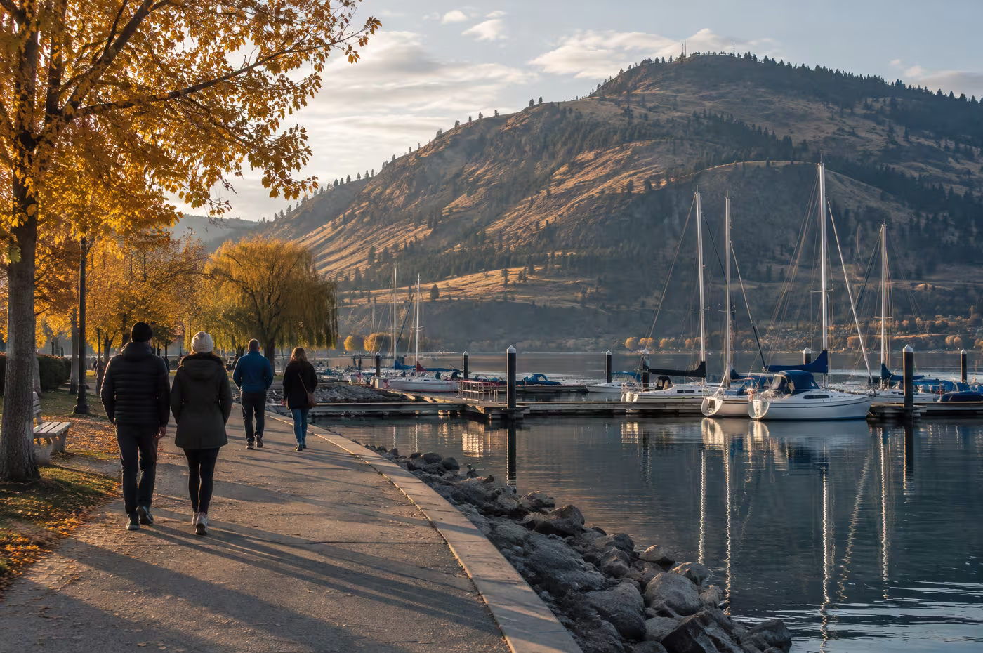 People walking the Kelowna waterfront past sailboats, Knox Mountain behind