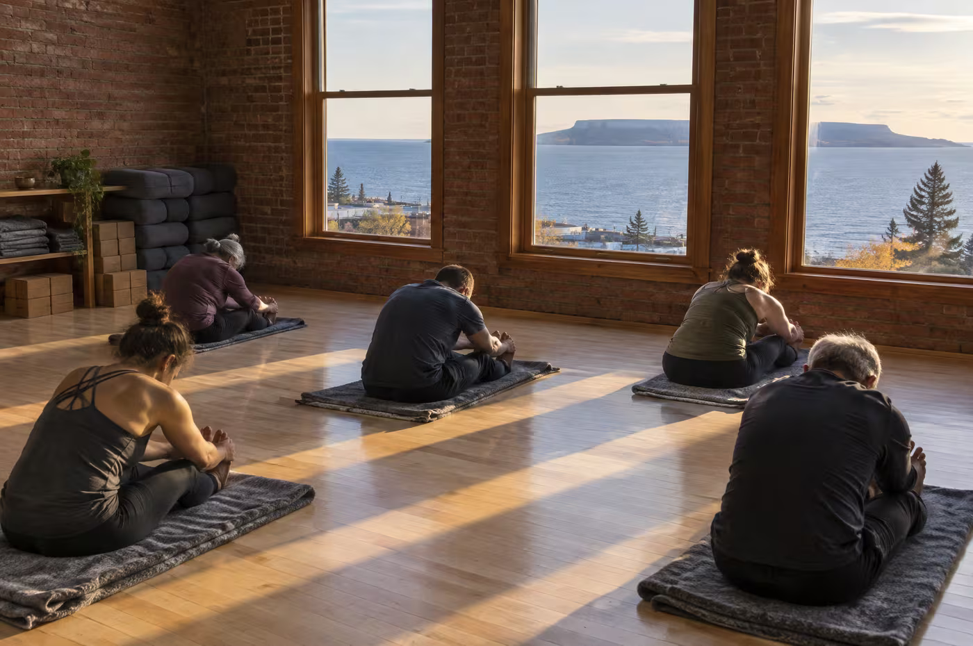 Students in a seated forward fold on wool blankets as low sun crosses a birch floor, the lake and the Sleeping Giant in the windows