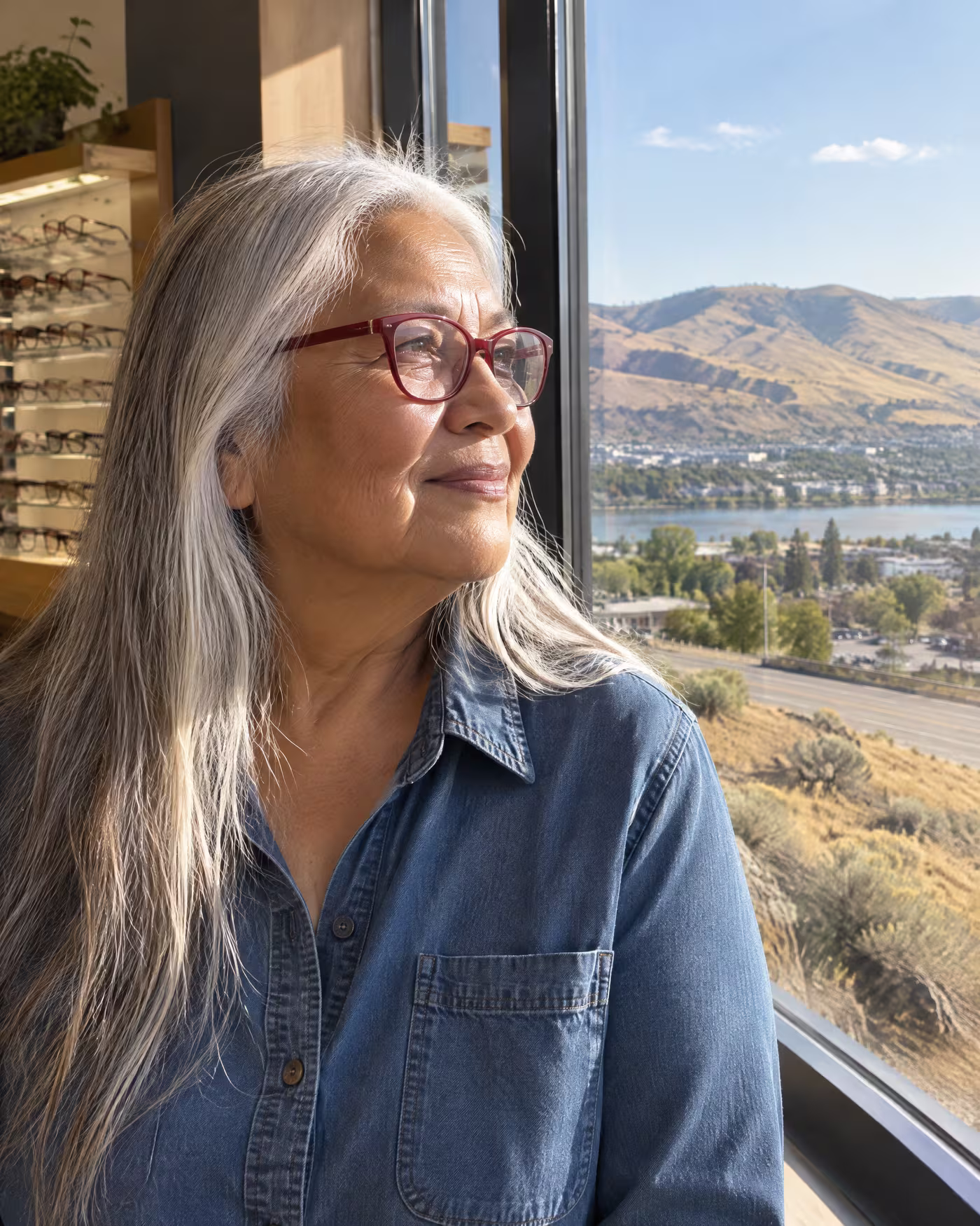 A woman with long silver hair in new deep red frames looking out toward dry hills