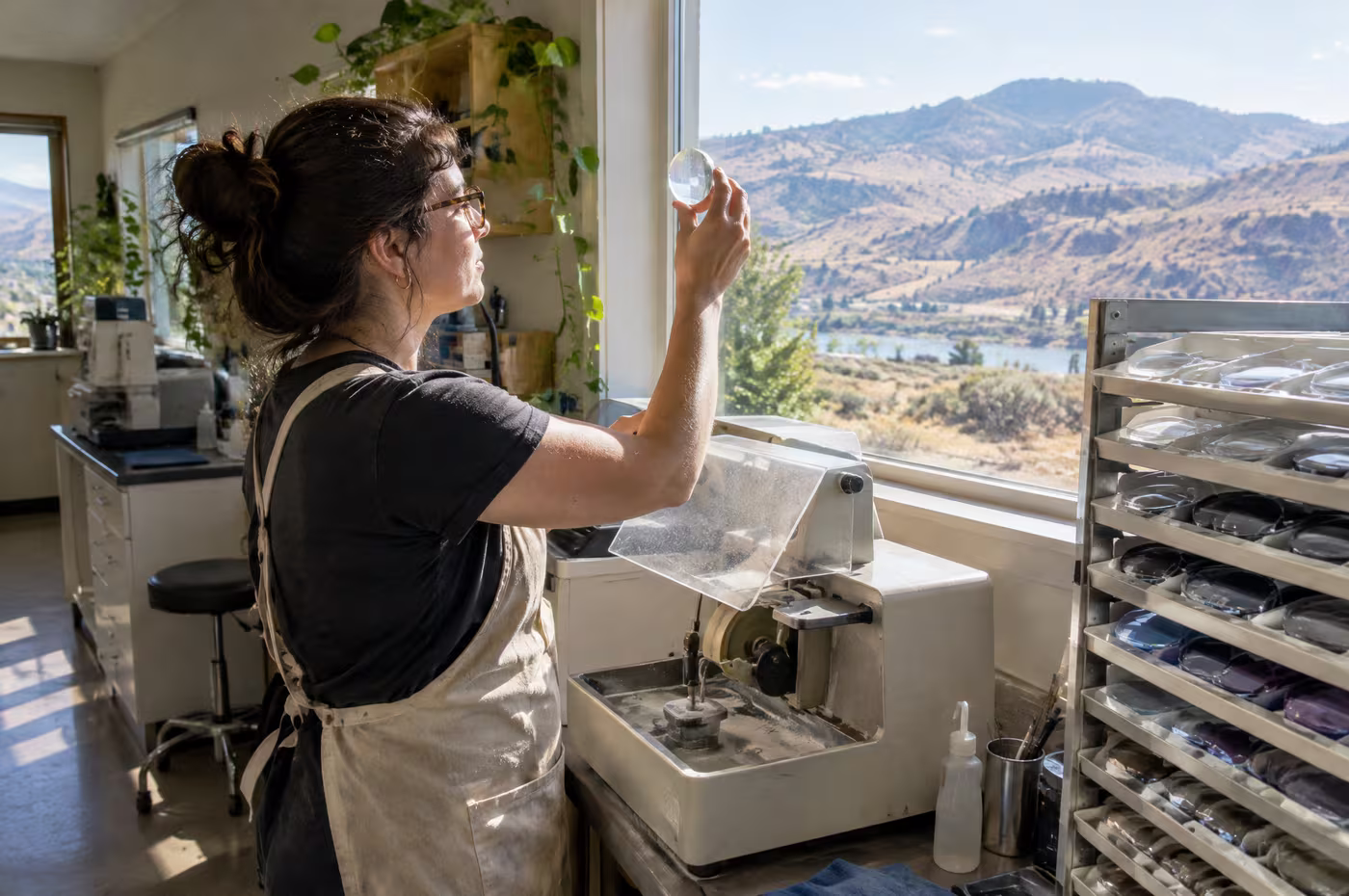 An optician in a canvas apron holding a freshly cut lens up to the window light in the workroom
