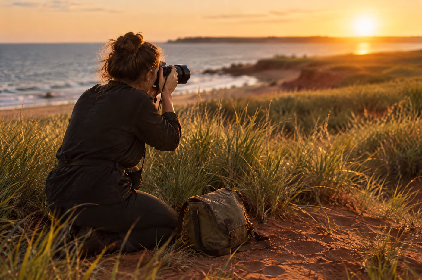 A photographer kneeling in dune grass with a camera raised, low sun over the red sand