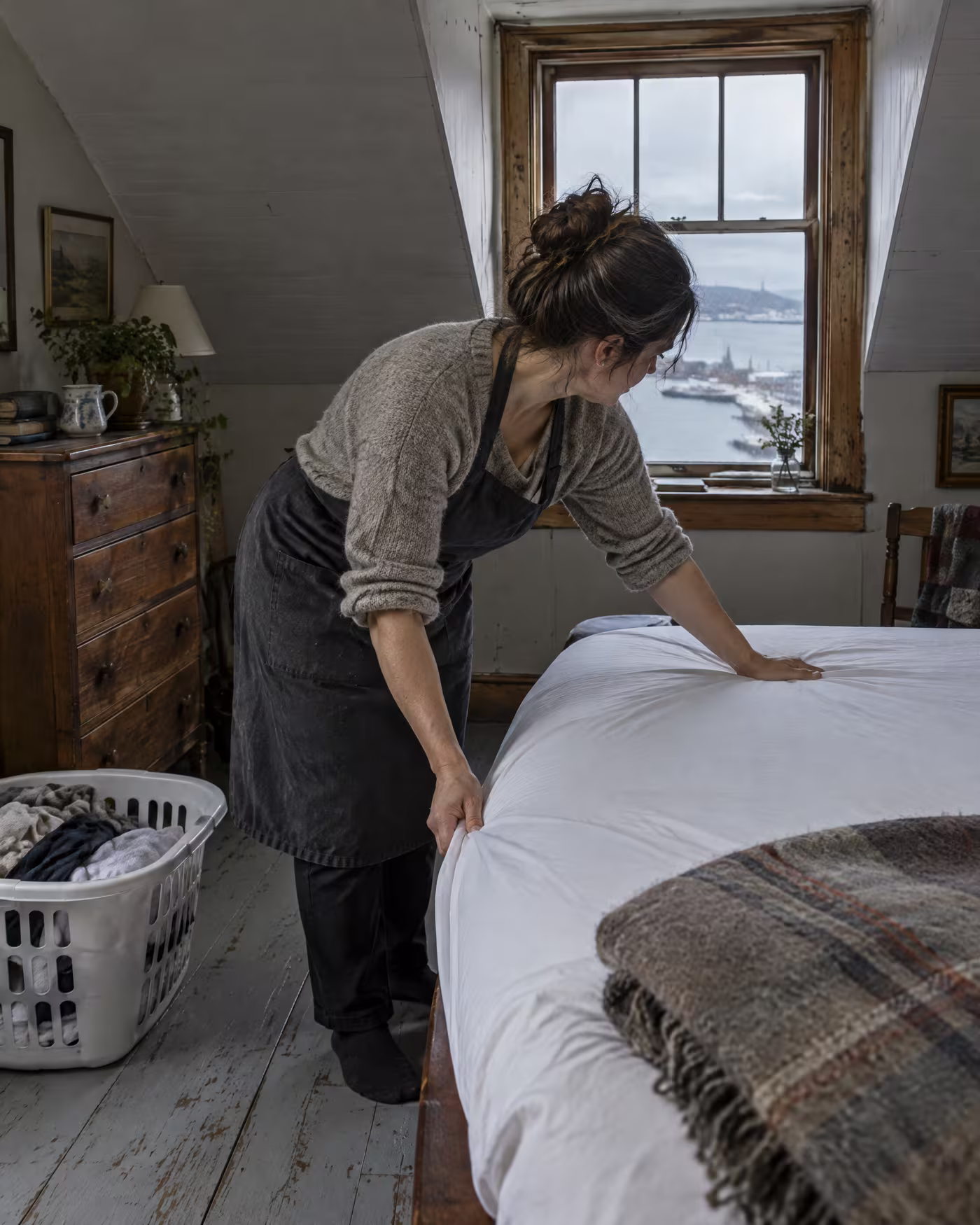 A cleaner smoothing a white fitted sheet onto a bed under the dormer window of an attic room