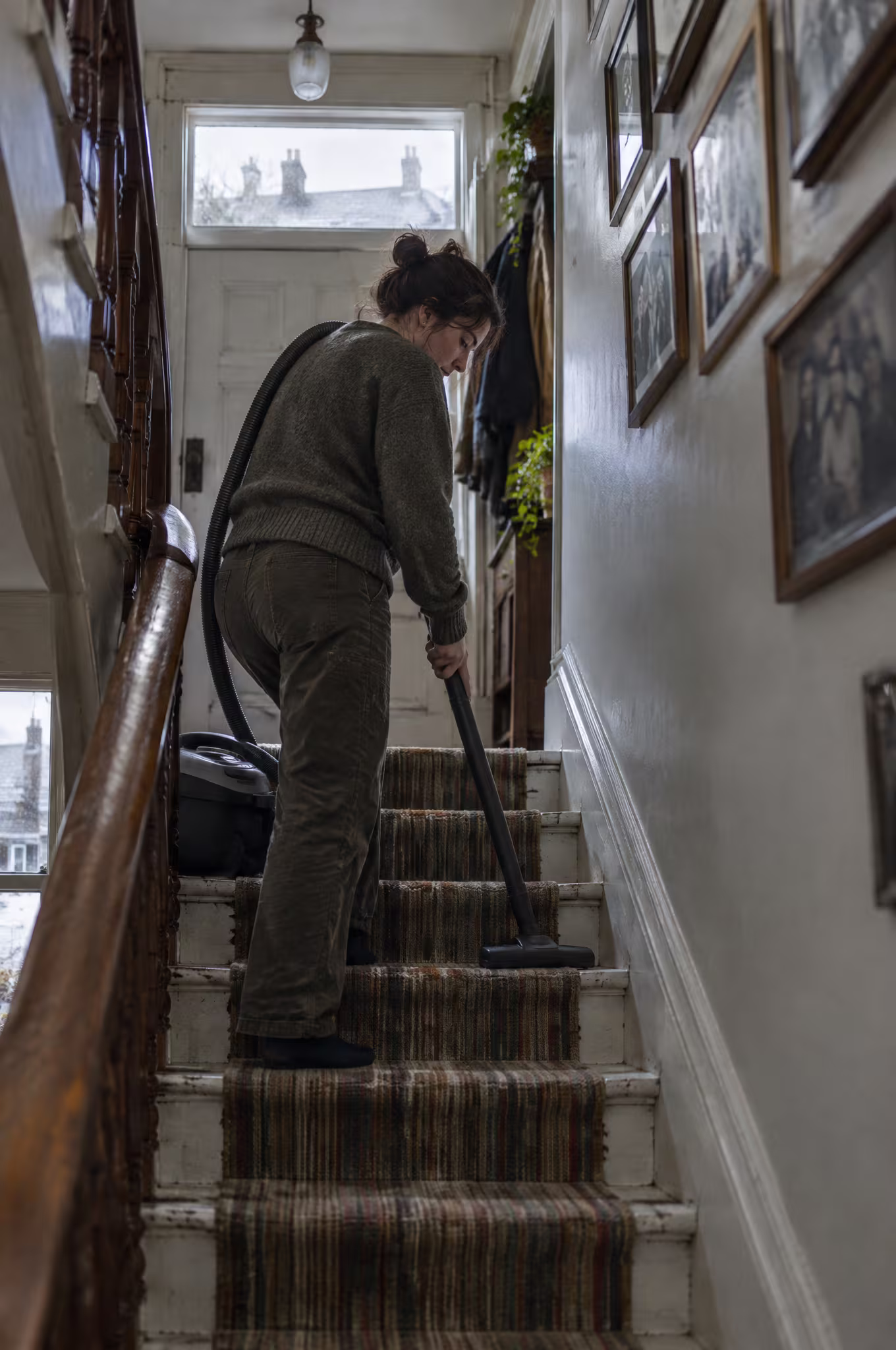 A cleaner vacuuming a steep painted staircase with a canister vacuum, light from the transom above the door
